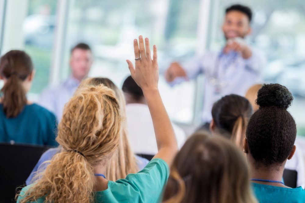 A woman raises her hand while a speaker points at her among a group of people being presented to